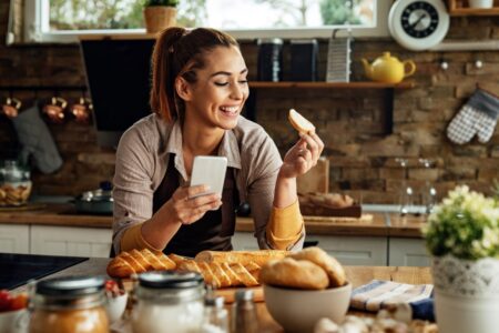 Young happy woman holding slice of bread while using smart phone and preparing food in the kitchen.